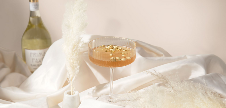 Dreamy image of non-alcoholic cocktail in a coupe glass garnished with jasmine buds. Setting is a cream fabric with white pampas grass and a non-alcoholic champagne bottle in the background. 